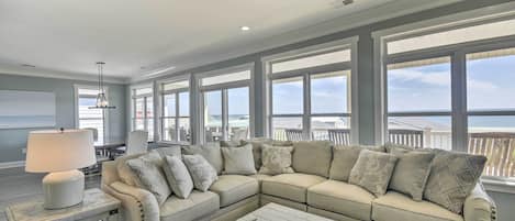 Amplia sala de estar con un sofá seccional beige grande, una mesa de centro y una mesita de noche con una lámpara. Ventanas grandes ofrecen vista a la playa.