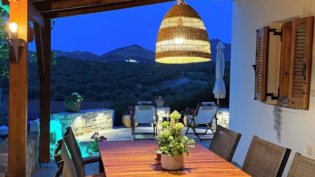 Un espace extérieur aménagé pour les repas, avec une table et des chaises en bois, un luminaire suspendu et une vue sur les montagnes en arrière-plan.