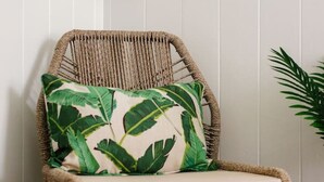 Una silla de mimbre con cojín decorado con un diseño de hojas verdes, ubicada frente a una pared blanca con paneles.