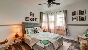 Un dormitorio con cama, ventilador de techo, mesita de noche, lámpara, silla y ventana con cortinas.