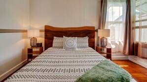 Ein Schlafzimmer mit einem hölzernen Kopfteil, gemusterten Betttextilien, zwei Nachttischlampen und einem mit Vorhängen versehenen Fenster.