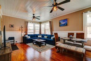 A cozy living room with a blue sofa, a coffee table, and a dining table with chairs. There are ceiling fans, a painting on the wall, and large windows with curtains. - NEW - Paddler`s Inn 3BR Hilo (Hilo)