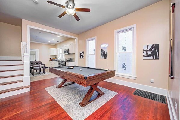 A pool table in a room with hardwood floors, a ceiling fan, and a kitchen in the background. - Spacious and Fun 5BR w/ Pool Table and more! (Indianapolis)