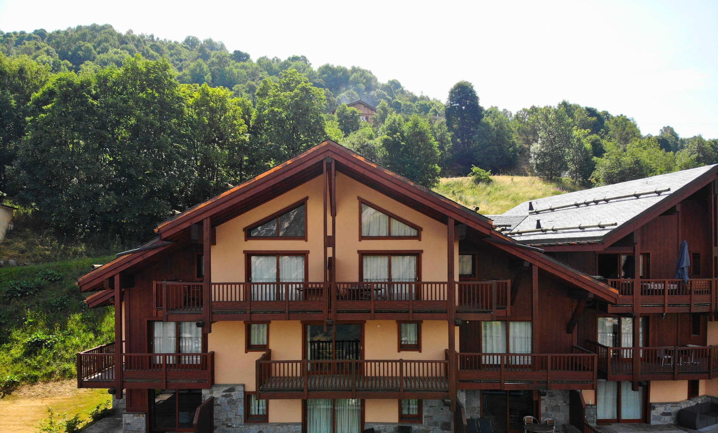 A multi-story chalet with balconies, stone foundation, and a view of a forested hillside.