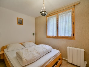 Een slaapkamer met een houten bed, wit beddengoed, een raam met kantgordijnen, een radiator en een klein schilderij aan de muur.