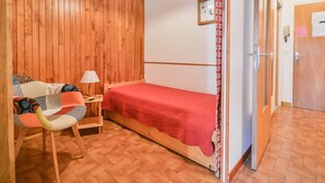 Ein kleiner Raum mit Holzwand, einem Bett mit roter Decke, einem Stuhl, einem Schreibtisch mit Lampe und einer Tür.