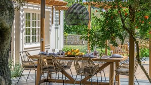 An outdoor dining area with a wooden table and wicker chairs, surrounded by greenery and a pergola. - Leeda's Cottages (Zakynthos)