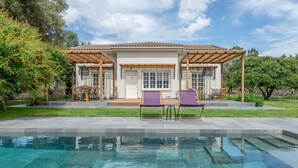 A poolside area with a pergola, two lounge chairs, and a house with a tiled roof. - Leeda's Cottages (Zakynthos)