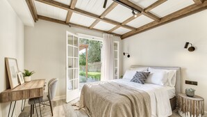 A bedroom with a large bed, a desk, and a view of a garden through an open door. - Leeda's Cottages (Zakynthos)