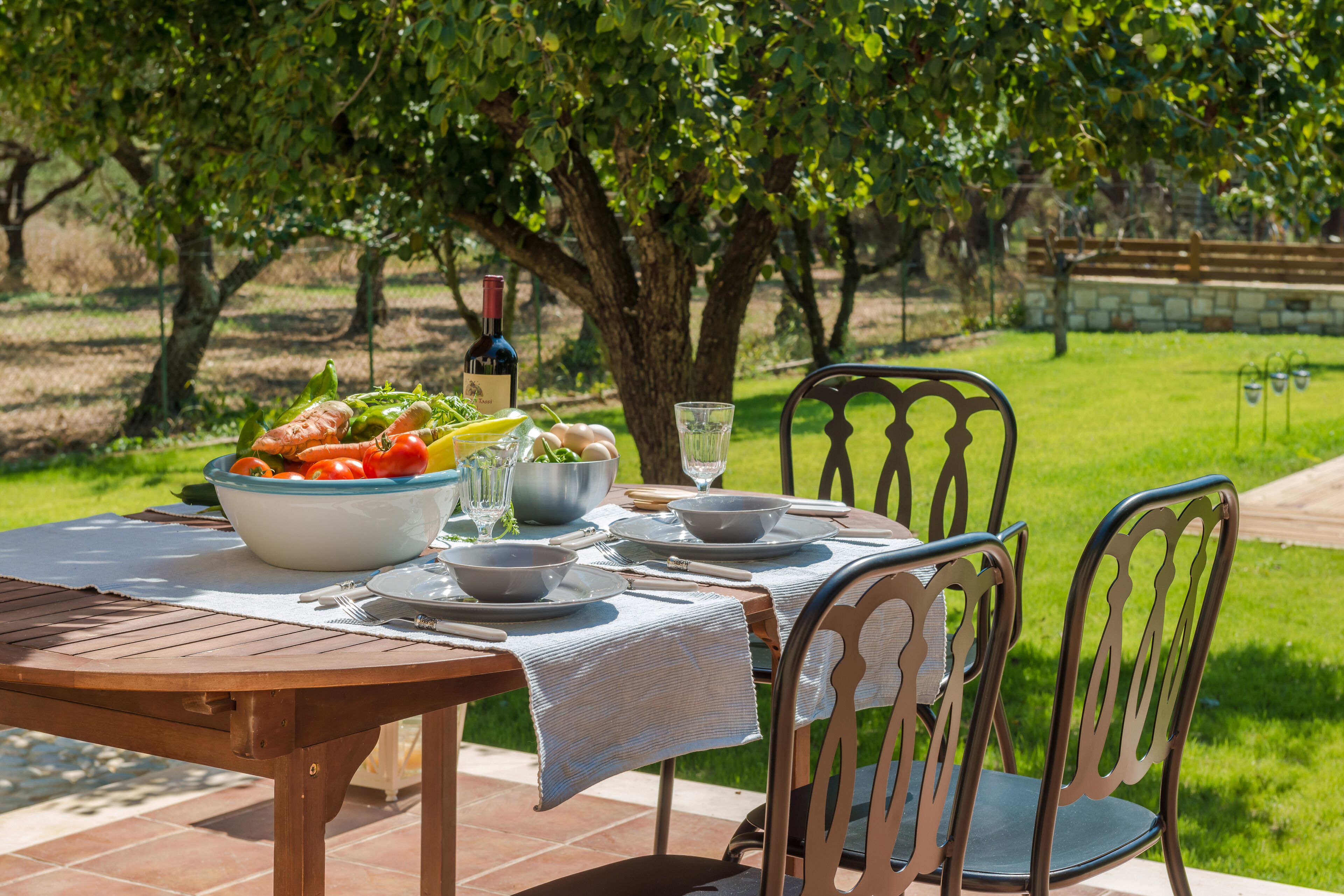 An outdoor dining table set with a bowl of vegetables, a wine bottle, and glasses, surrounded by chairs and a tree.