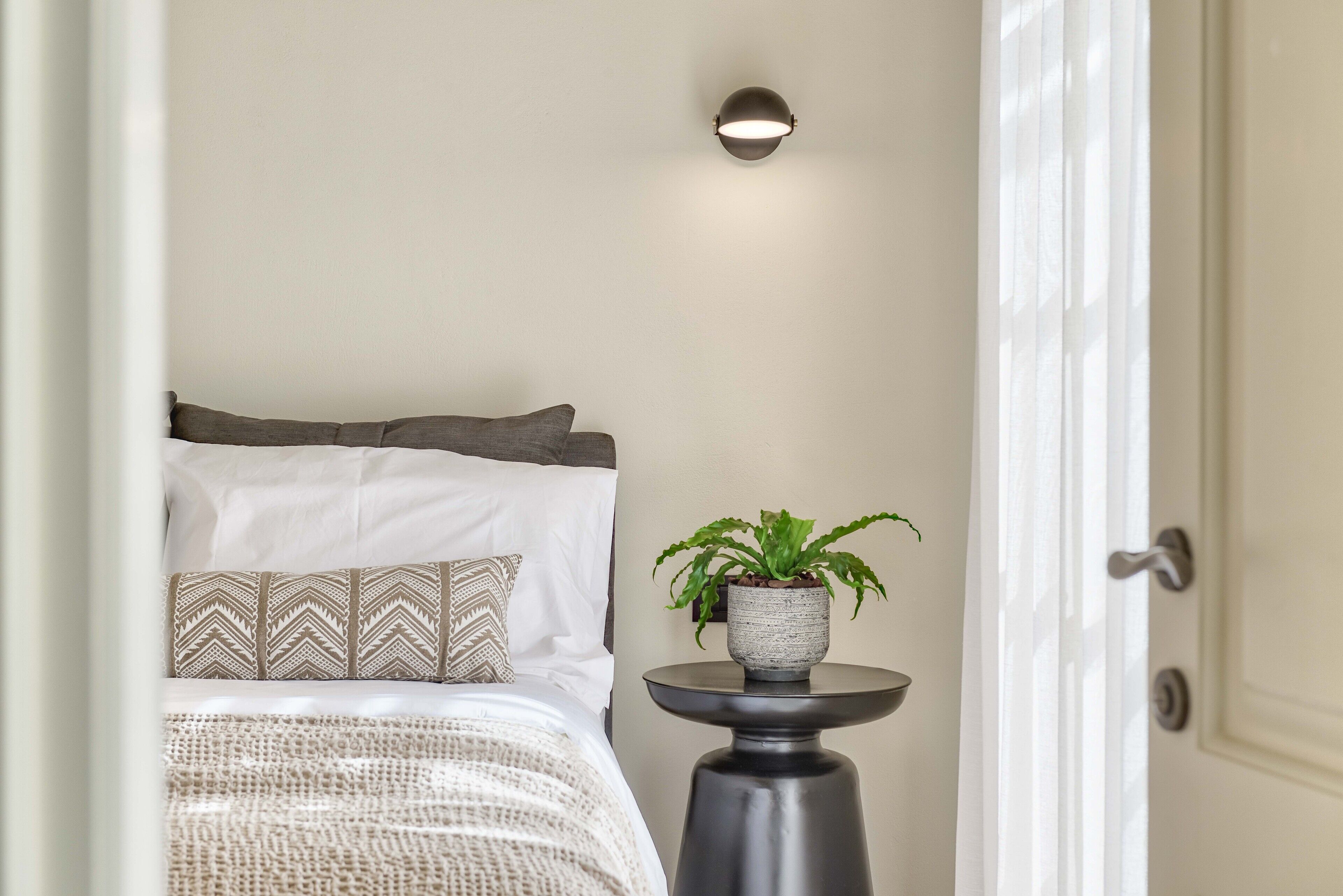 A bedroom with a bed, pillows, a bedside table with a plant, and a wall-mounted light fixture.