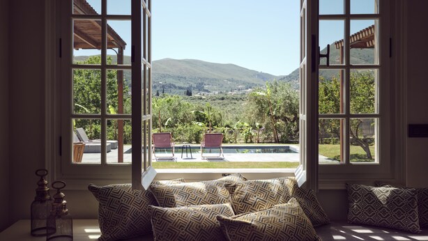 A room with a large window overlooking a pool and mountains. There are several patterned cushions on a bench. - Leeda's Cottages (Zakynthos)