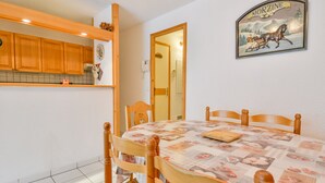 Uma sala de jantar com mesa posta para quatro pessoas, cadeiras de madeira, área de cozinha com armários de madeira e um quadro pendurado na parede.