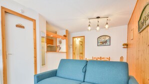 Sala de estar com sofá azul, paredes de madeira, cozinha e mesa de jantar com cadeiras.