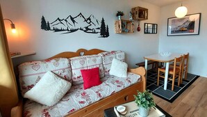 A cozy living room with a wooden sofa, a small table, a wall-mounted shelf, and a dining area with a table and chairs. - Cozy 2-room apartment in La Plagne, near the slopes (La Plagne-Tarentaise)