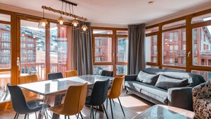 A modern living room with a glass dining table, comfortable seating, and large windows offering a view of a snowy landscape. - 3-room apartment with balcony, WIFI, parking and pool access, Les Arcs 1800 (Les Arcs 1800)
