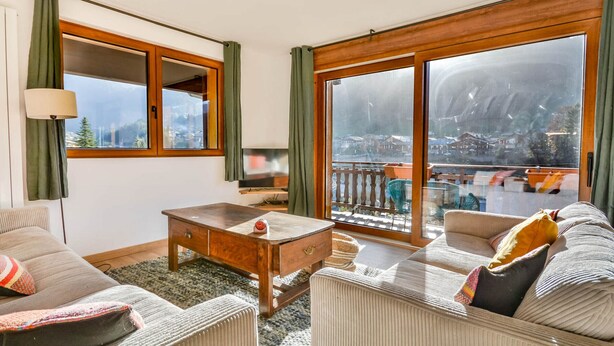 A living room with a sofa, a coffee table, and a TV. There are large windows with curtains, and a balcony with outdoor furniture. - Stunning renovated T5, bright, sleeps 10 in Morzine (Morzine)