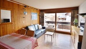A room with a wooden paneled wall, a sofa, a dining table with a red and white checkered tablecloth, a small coffee table, and a view of a balcony with outdoor furniture. - Studio in Morzine center, 4 pers, balcony, WIFI (Morzine)