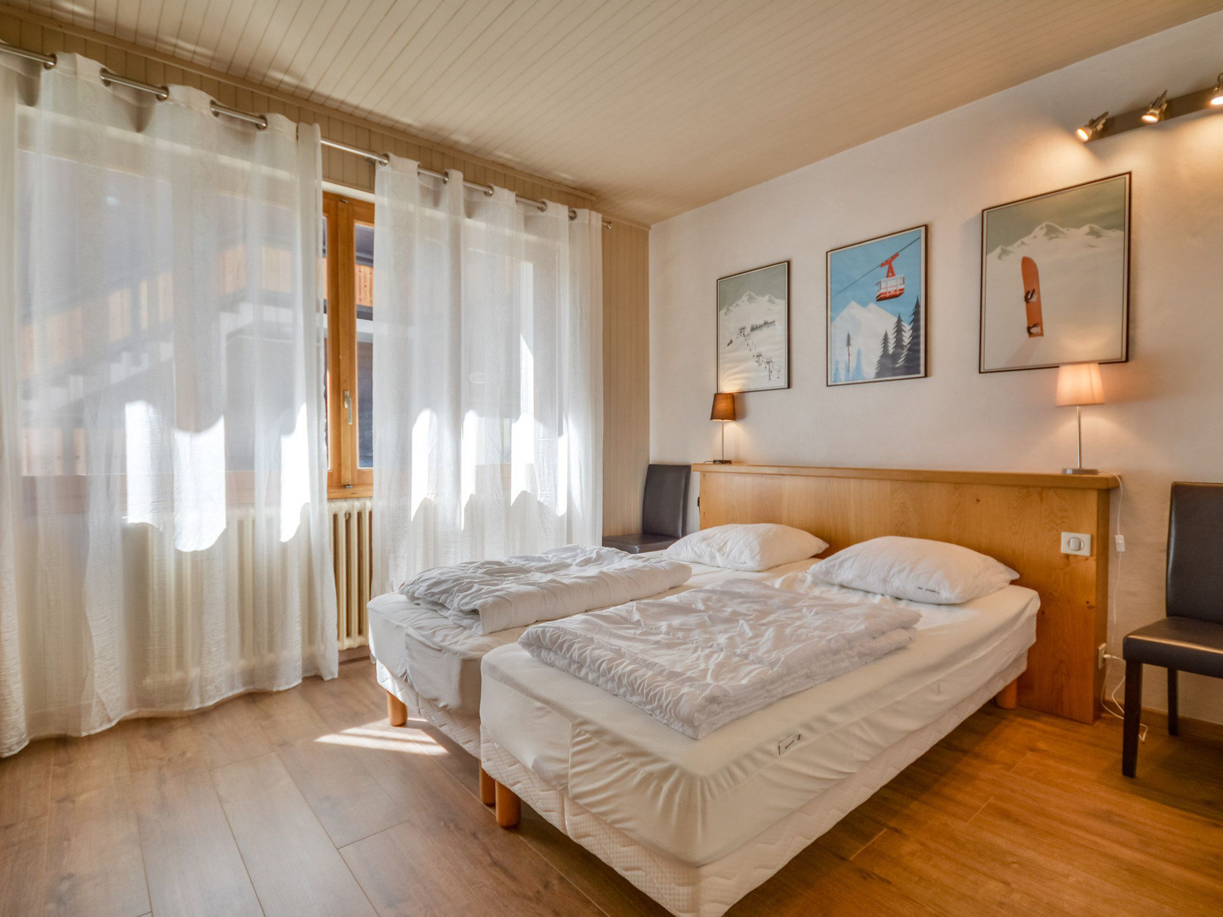 A hotel room with two beds, a wooden headboard, a window with sheer curtains, and wall-mounted lamps.