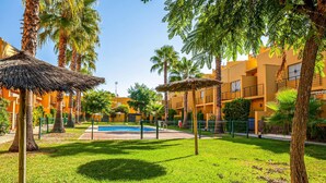 A pool area with a thatched umbrella, palm trees, and a building with balconies in the background. - 2 bedroom amazing home in Isla Cristina (Isla Cristina)