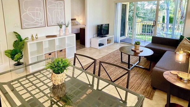 A modern living room with a glass dining table, a large woven ceiling light, a sofa, a TV, and a balcony with a view of greenery. - 260D - Boca Retreat w/ Pool, Parking, Near Evert Tennis & Shops (Boca Raton)