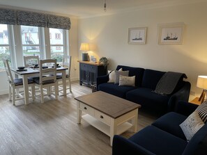 A living room with a dark blue sofa, a wooden coffee table, white chairs, a dining table set for two, and a cabinet. - Beautiful coastal apartment (Cellardyke)
