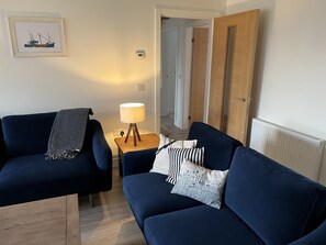 A modern living room with a dark blue sofa, a small wooden side table with a lamp, and a framed picture on the wall. - Beautiful coastal apartment (Cellardyke)