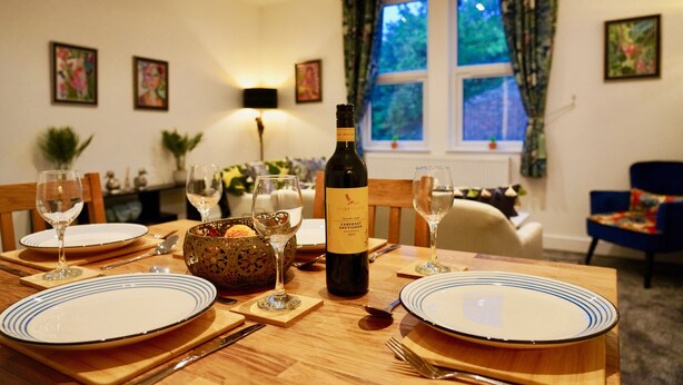 A dining table set with plates, wine glasses, and a bottle of wine. There are chairs around the table, and a window with curtains in the background. - Funky 2 Bed Apartment in the Heart of Historic Rochester (Rochester)