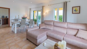 Een moderne woonkamer met een beige modulaire bank, een eethoek met een witte tafel en stoelen, en een keuken op de achtergrond.