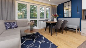 Una sala moderna con sofá, mesa de comedor y sillas. Hay ventanas amplias con cortinas, un florero con flores y una pared azul.