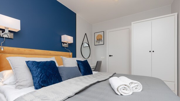 Una cama bien hecha con colcha gris y blanca, almohadas azules y blancas, y toallas blancas enrolladas sobre una colcha gris.