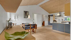 En rummelig opholdsstue med træloft, spisebord med stole, et køkken med blå skabe og et stort vindue.