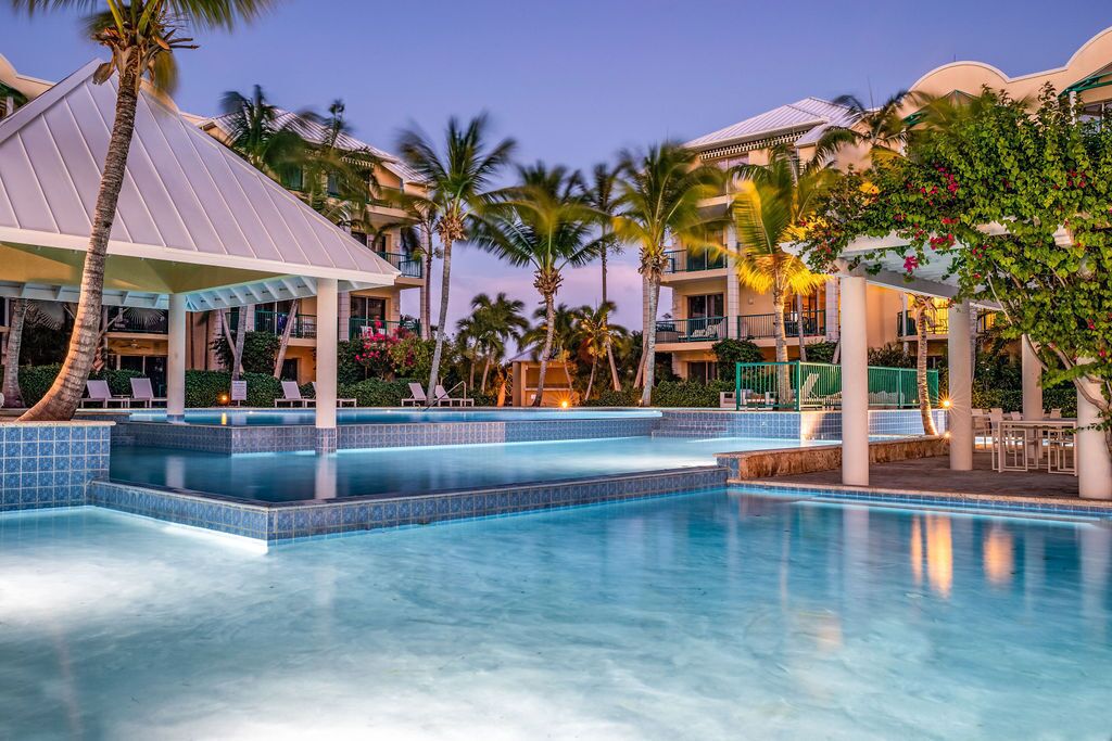 Um hotel com piscina, palmeiras e área de estar coberta.