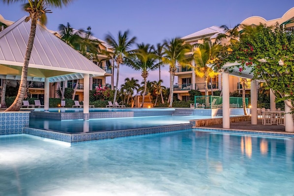 A hotel with a swimming pool, palm trees, and a covered seating area.