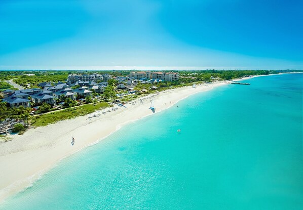 An aerial view of a coastal resort with beachfront properties, a sandy beach, and clear turquoise waters.
