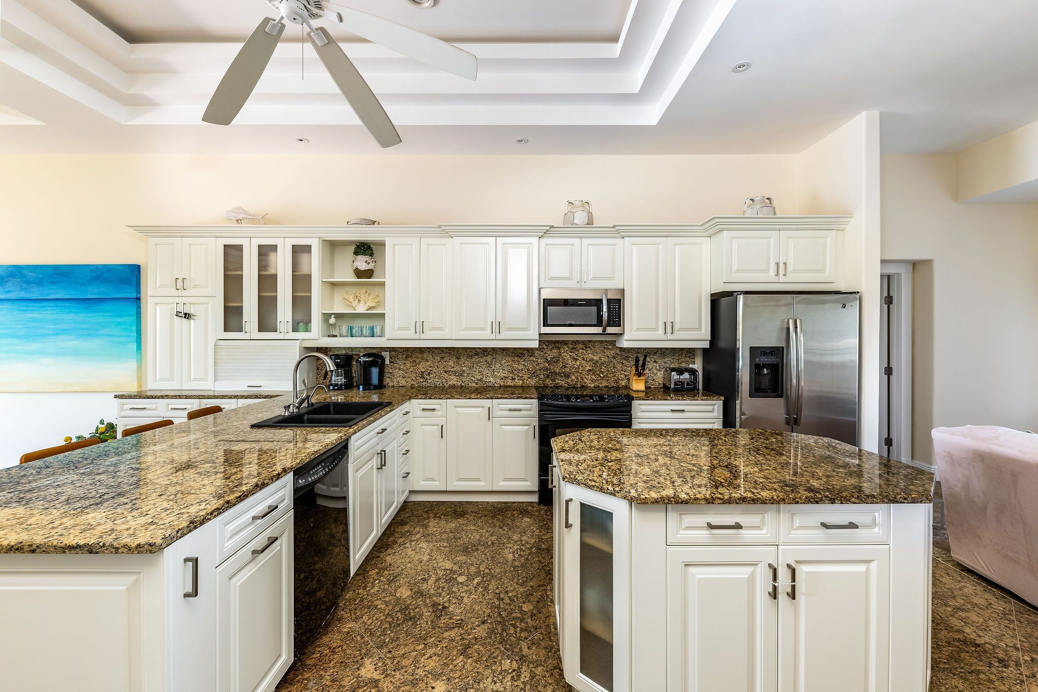 Una cocina moderna con muebles blancos, encimeras de granito, un refrigerador de acero inoxidable y un ventilador de techo.