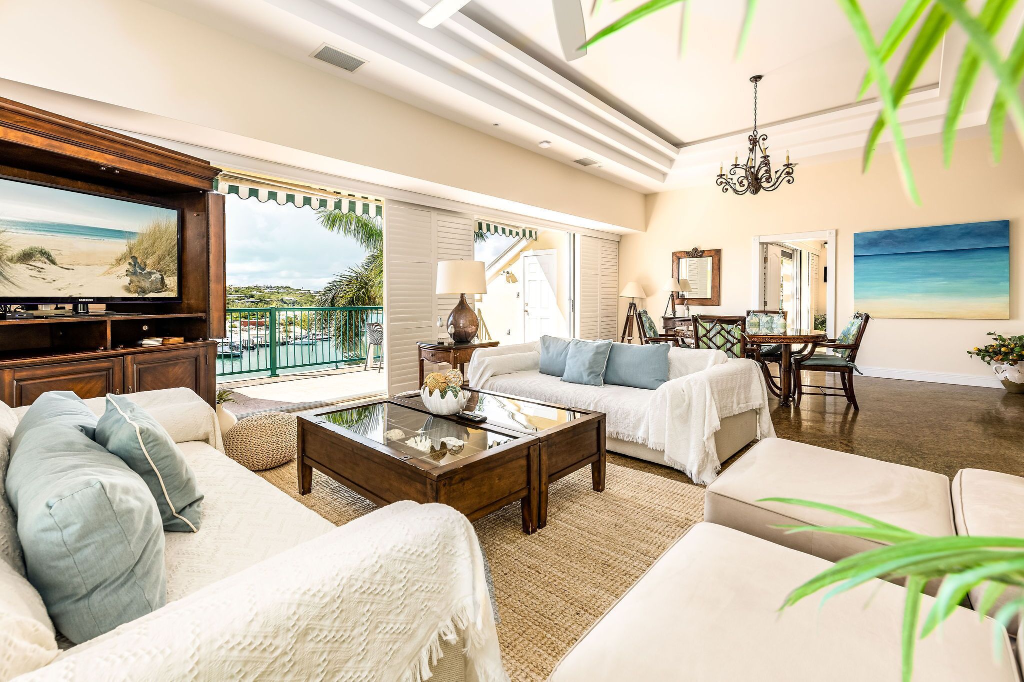 A spacious living room with a large flat-screen TV, comfortable seating, a dining area, and a balcony with a view.