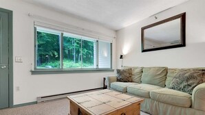 Une pièce avec une grande fenêtre, une commode en bois, un canapé de couleur claire et un miroir au mur.
