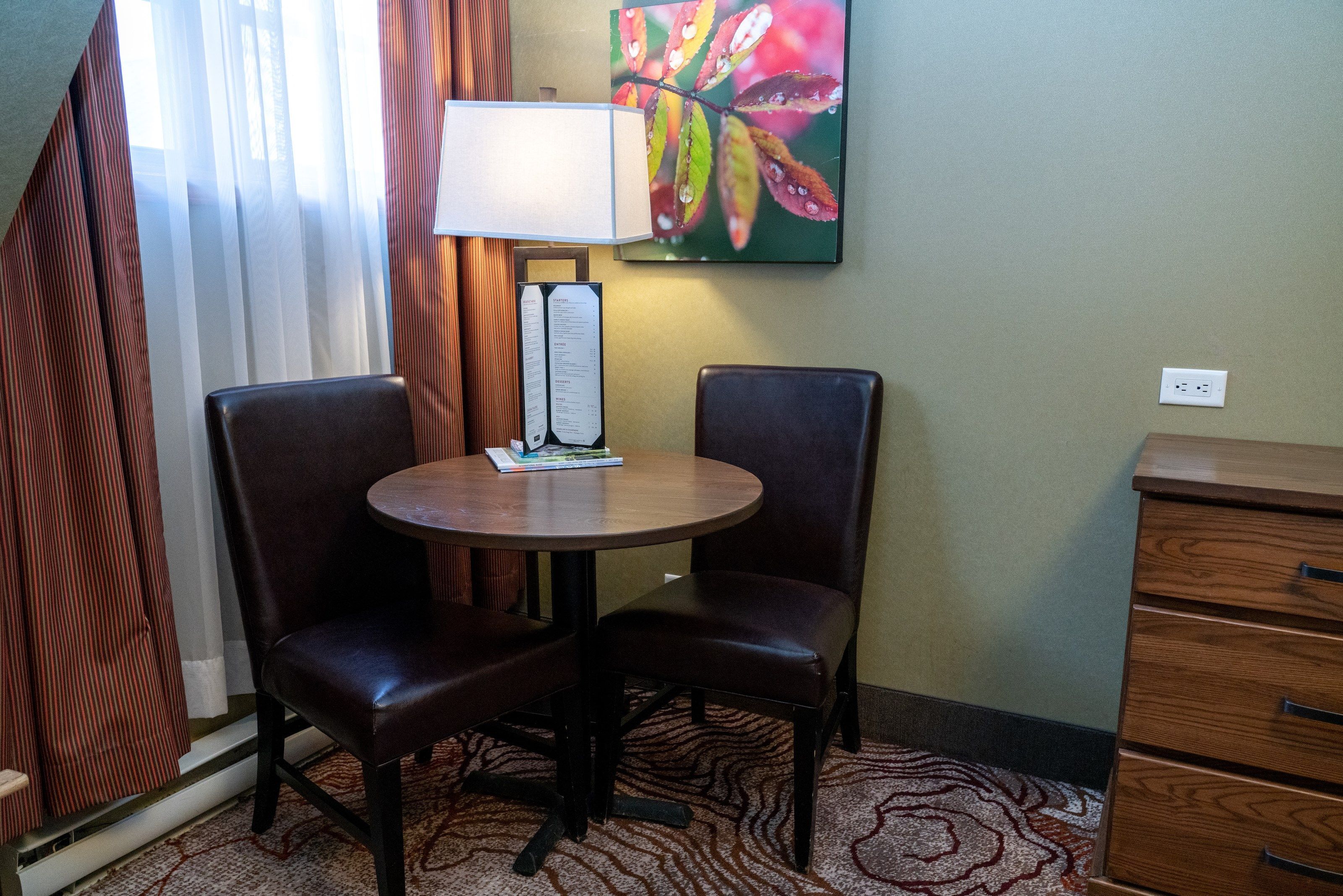 Een hotelkamer met een ronde tafel, twee stoelen, een lamp, een schilderij en een houten dressoir.