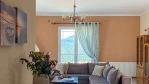 A living room with a sectional sofa, a coffee table, and a vase with flowers. There is a window with curtains, a chandelier, and a cabinet. - Fotis Apartment (Kefalonia)