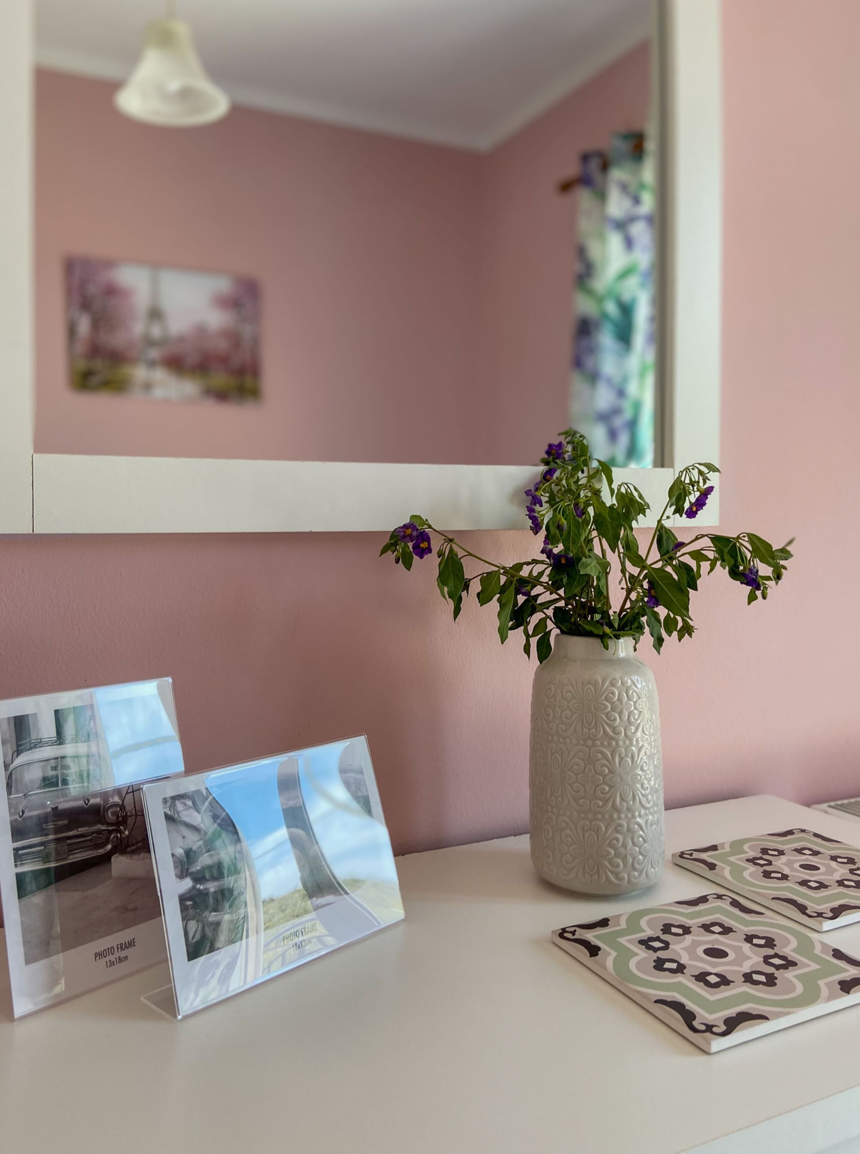 A white table with a white vase containing purple flowers, a photo frame, and patterned tiles.