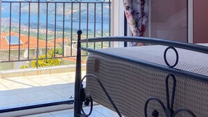 A balcony with a metal railing, a bed with a white mattress, and a view of a mountainous landscape. - Fotis Apartment (Kefalonia)