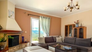 A living room with a fireplace, a sofa set, a coffee table, and a view of the outdoors. - Fotis Apartment (Kefalonia)