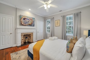 A bedroom with a fireplace, a bed with white linens and a yellow throw, a ceiling fan, and a large abstract painting.
