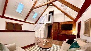 A spacious loft with a high ceiling, exposed wooden beams, a staircase, a cozy seating area with a coffee table, and a flat-screen TV mounted on the wall. - New! The Sanctuary House w/mountain view sleeps 14 (Medicine Park)