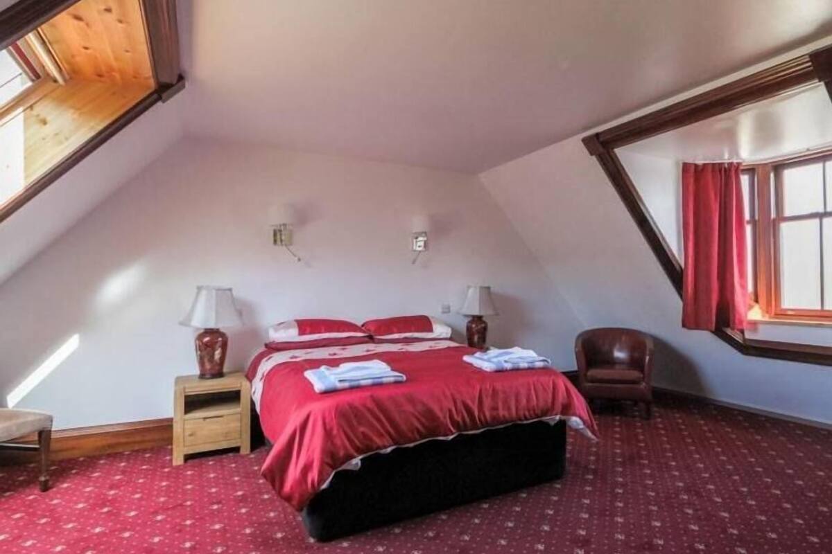 A bedroom with a sloped ceiling, a bed with red bedding, two bedside lamps, a wooden nightstand, a chair, and a window with red curtains.