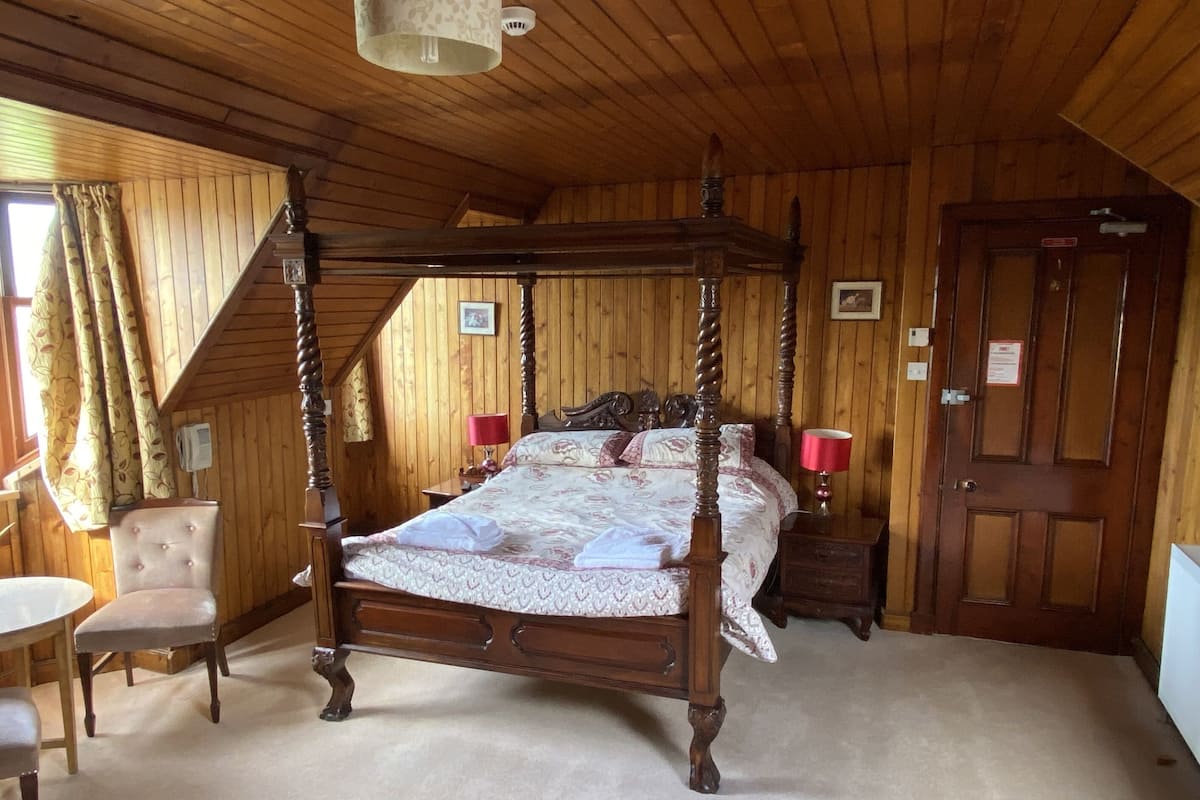 A four-poster bed with a floral bedspread, a wooden chair, a small table, and a door with a sign.