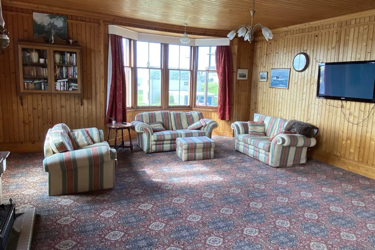 A cozy living room with striped sofas, a wooden ceiling, a large window, and a television on the wall.