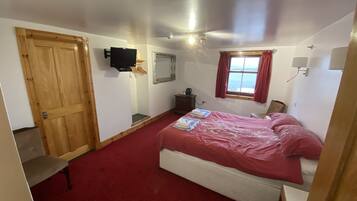 Un dormitorio con alfombra roja, cama con ropa de cama rosa, puerta de madera, televisor montado en la pared, una mesita de noche pequeña y una ventana con cortinas.