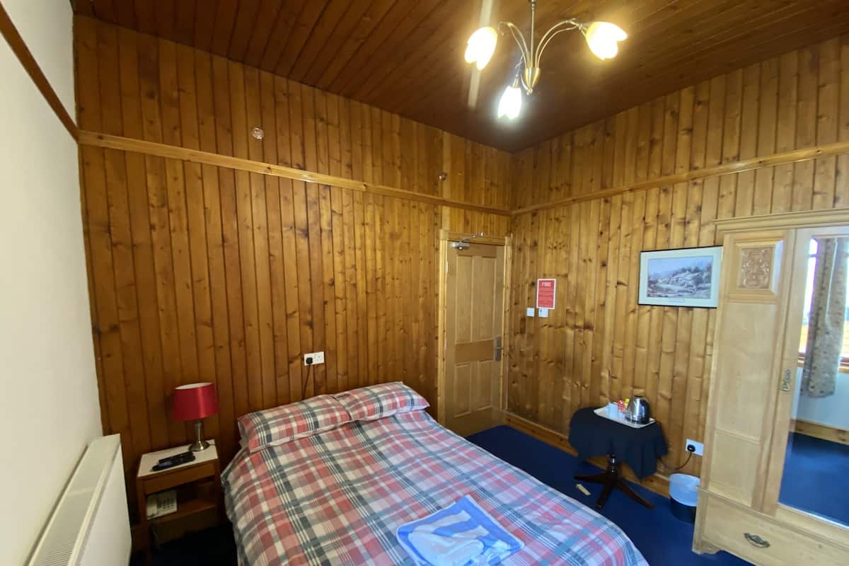 A wooden-paneled room with a bed, a small table with a lamp, a radiator, and a wardrobe.